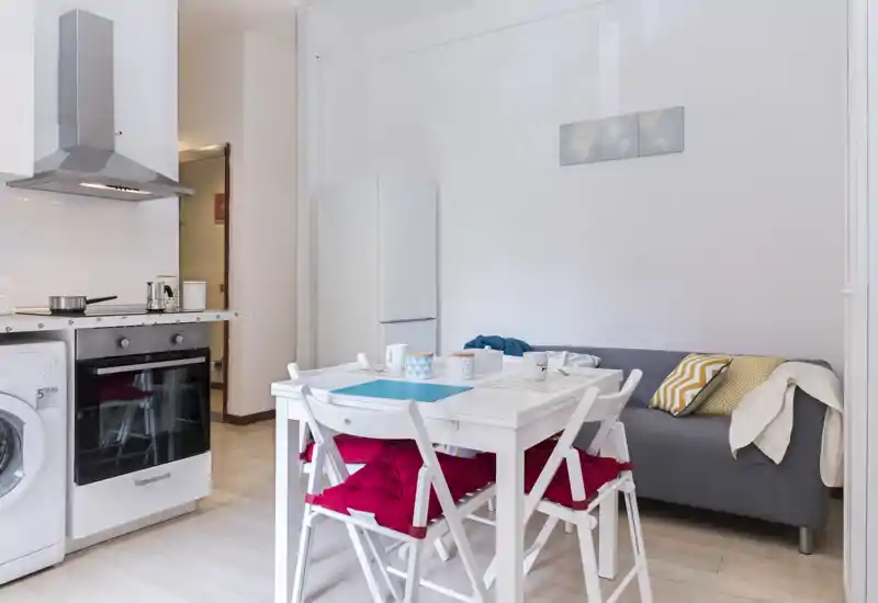 Bright open kitchen with a compact dining table and a small sofa; modern appliances, a hood and built-in oven, plus cozy cushions and table settings for four.