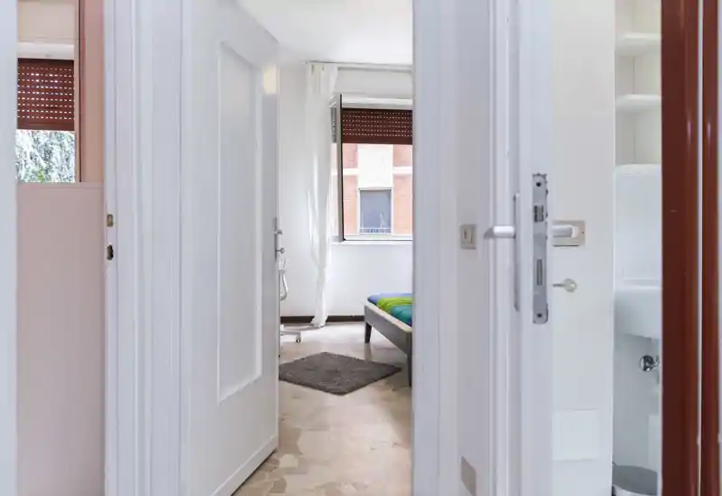 Bright, simply furnished bedroom seen through an open doorway with a visible bed, small rug and natural light from a window.