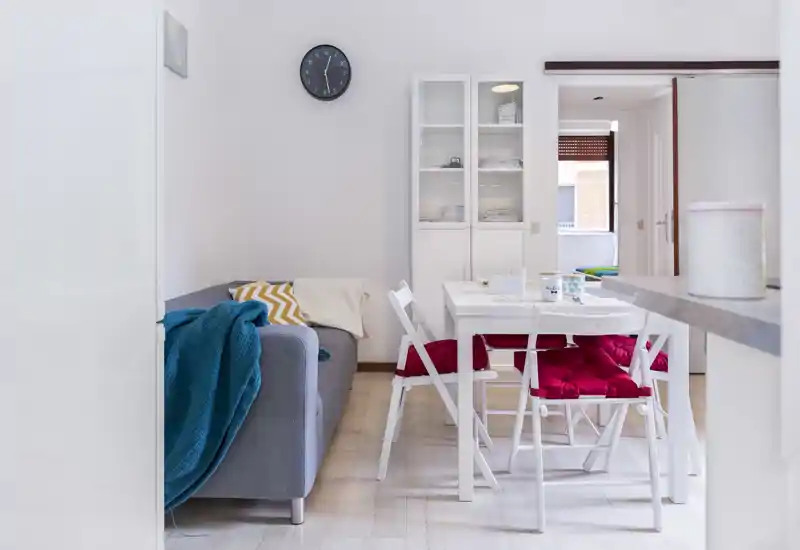 Bright, well-staged living area with a compact dining table, cozy sofa with throws and colorful cushions, and a storage cabinet — ideal for a small apartment.