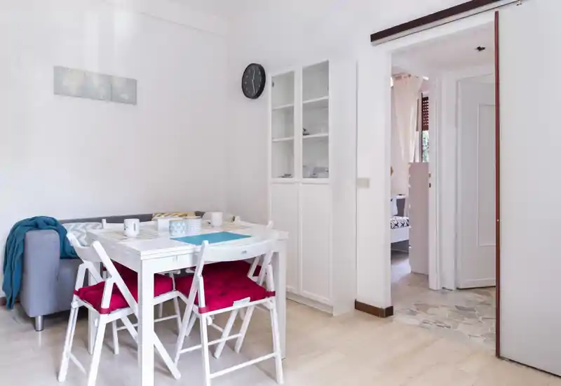 Bright, compact dining nook with a white table, four chairs with red cushions and a small sofa beside it — ideal for cozy meals in a modern apartment.