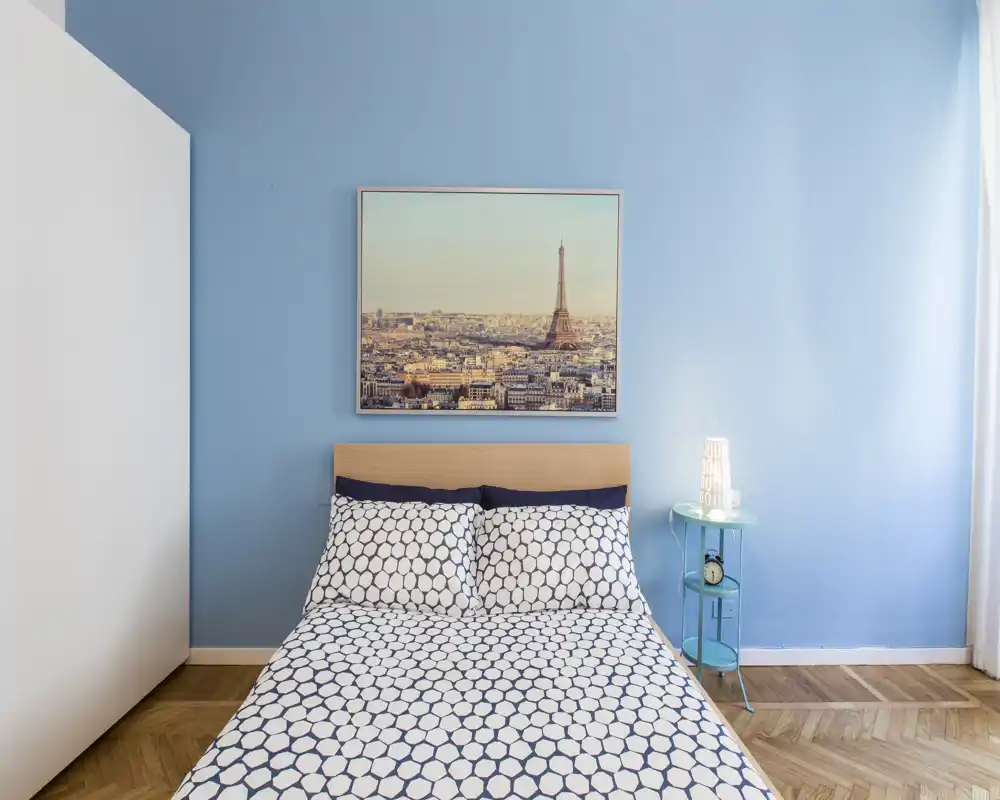 Bright, modern bedroom with a double bed centered against a soft blue wall, patterned bedding, a framed Paris print above the headboard and a small bedside table with a lamp.