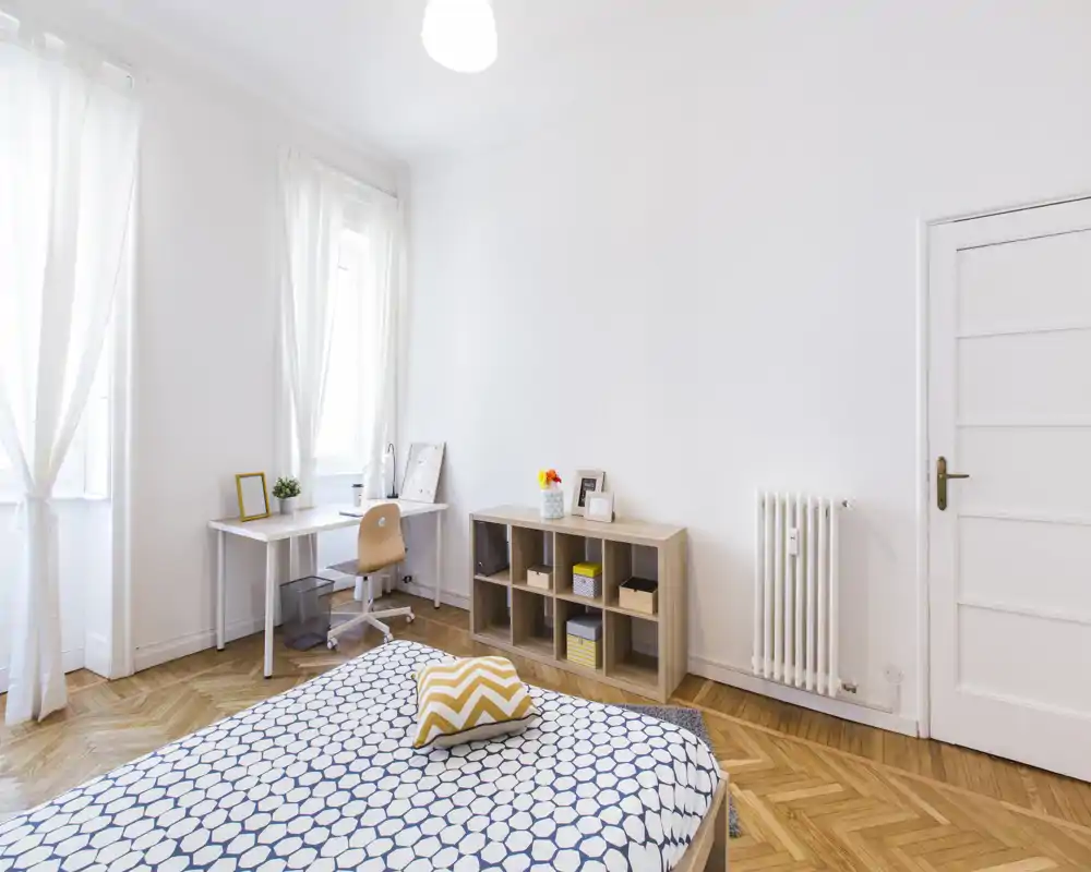 Bright, minimalist bedroom with a patterned duvet, decorative cushion and a compact desk by the window. Light wood flooring and open shelving create a calm, airy atmosphere ideal for restful stays.