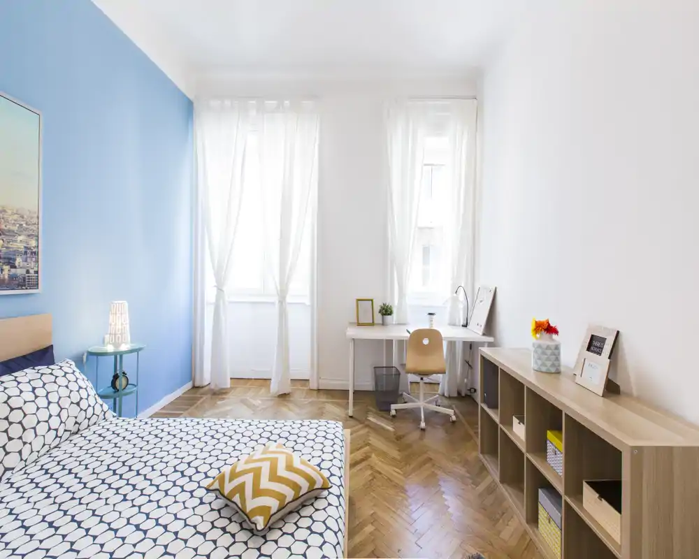 Bright, modern bedroom with patterned bedding, a bedside table and a small workspace by large windows; light hardwood floors and soft textiles create a welcoming atmosphere.