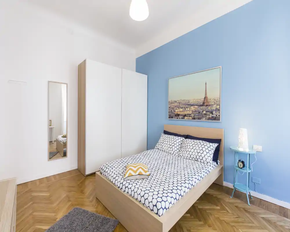 Sunny, modern bedroom with a double bed, patterned bedding, light wood frame and a blue accent wall featuring framed city artwork. The room includes a wardrobe, bedside table with lamp and a full-length mirror, styled with warm parquet flooring.