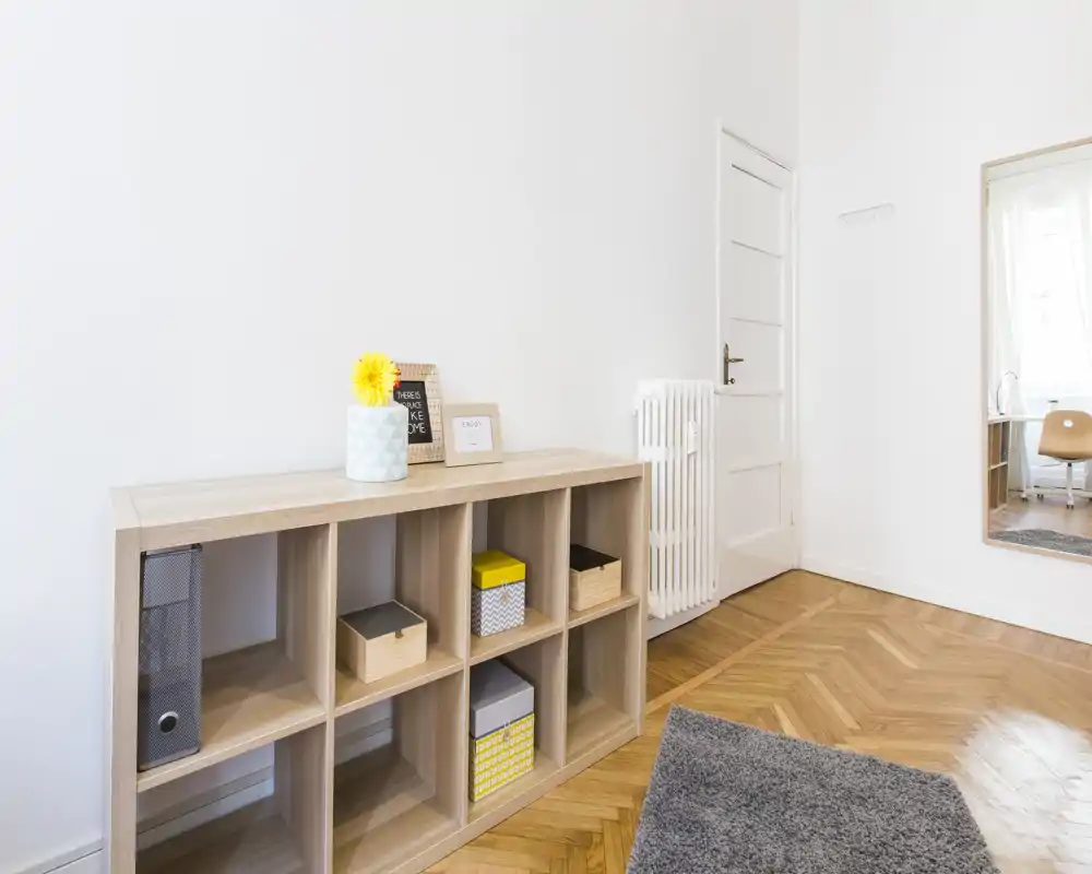 Bright, minimal living area with a light-wood cubby shelving, decorative boxes and a soft rug. Clean white walls and natural light create an airy, modern feel.