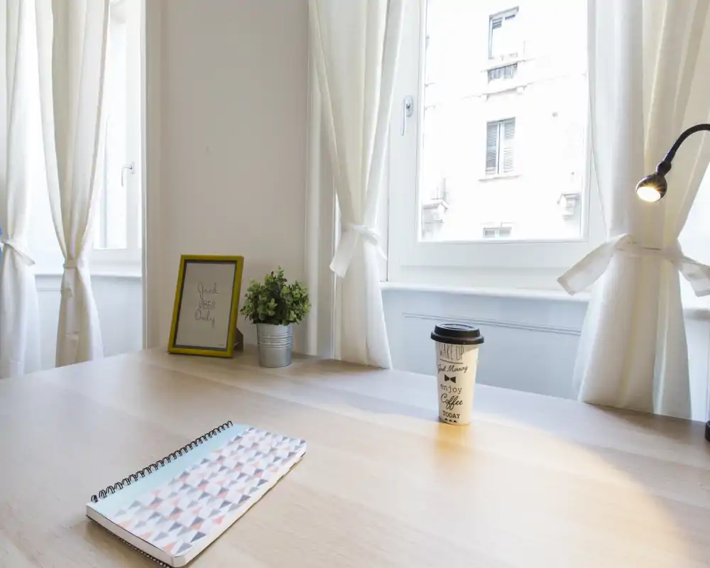 Bright corner workspace with a clean wooden table, potted plant and a notebook — naturally lit by large windows with soft white curtains.