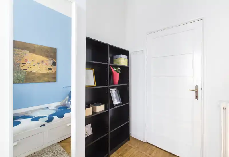 Chambre compacte et lumineuse vue à travers l'encadrement d'une porte : lit banquette blanc avec parure à motifs sur un mur bleu clair, tableau décoratif et étagère noire à proximité.