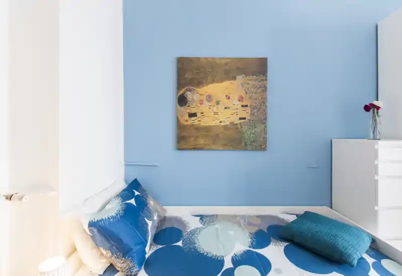 Compact, well-styled bedroom corner with a patterned blue bed, decorative pillows and a framed artwork on a pale blue wall — bright and cozy for a single or guest room.