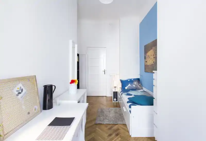 Bright, compact bedroom with a single daybed against a blue accent wall, wooden herringbone flooring and tidy workspace — ideal for a short-stay or student rental.