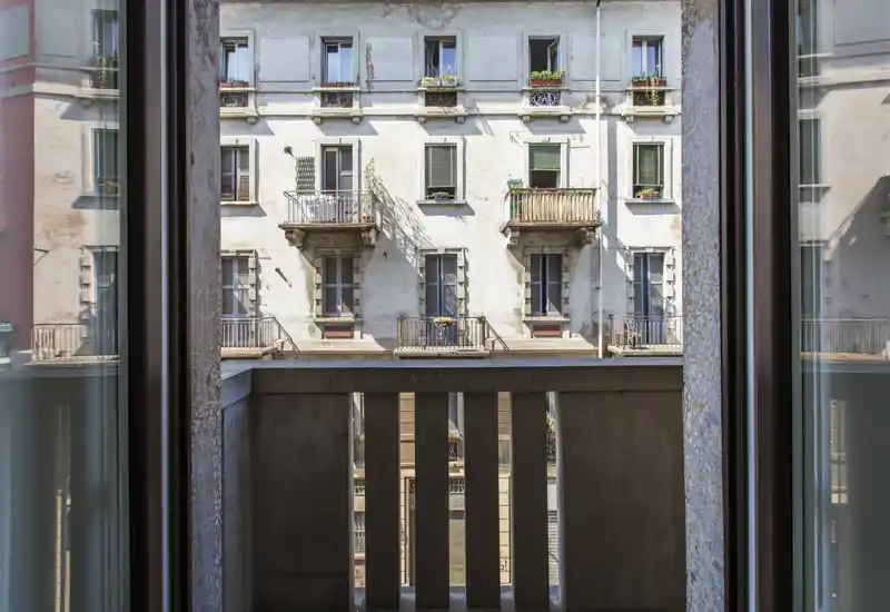 Petit balcon encadré par une fenêtre ouverte, vue sur la façade d’un immeuble voisin avec balcons et jardinières.