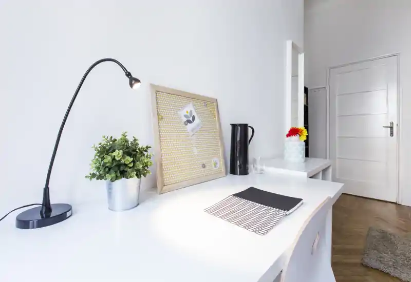 Coin salon lumineux et minimaliste avec bureau blanc, lampe articulée, plante en pot et panneau décoratif — propre et bien éclairé, idéal pour travailler ou lire.