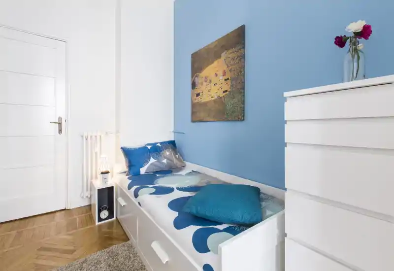 Small bright bedroom with a single daybed against a blue accent wall, patterned bedding, cushions and a white chest of drawers — cozy and well lit.