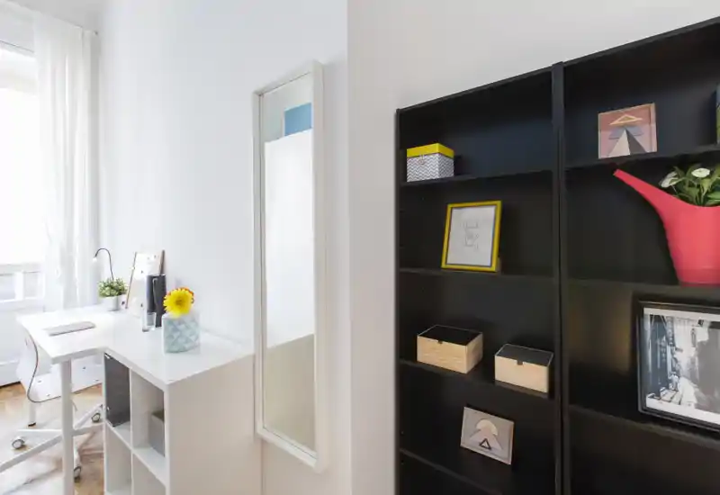 Espace de vie lumineux et moderne avec bureau blanc et étagères : coin travail ensoleillé à côté d'une grande bibliothèque noire, boîtes décoratives et plantes.