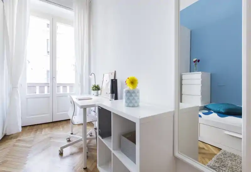 Coin de chambre lumineux et moderne avec lit partiellement visible, rangement blanc et petit bureau près de grandes fenêtres laissant entrer beaucoup de lumière naturelle.