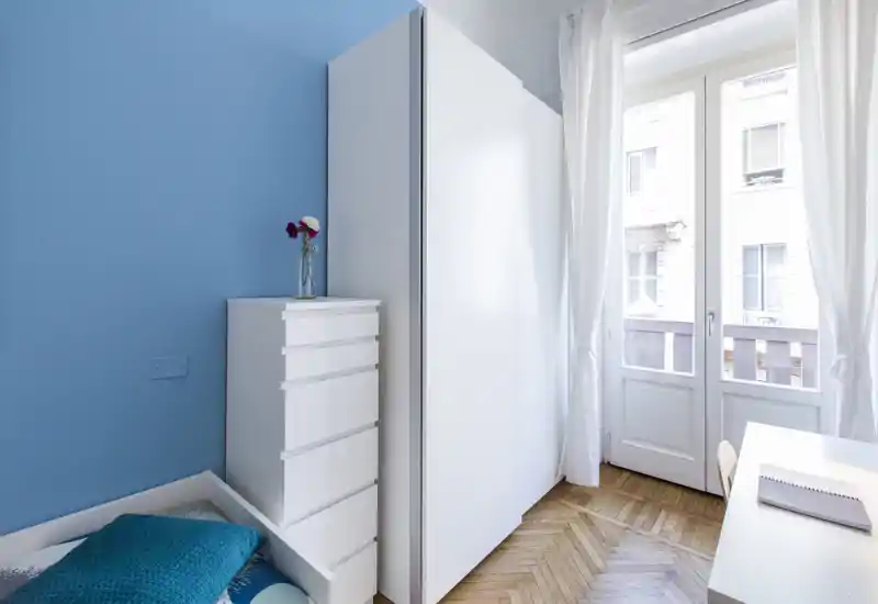 Bright, minimal bedroom corner with a white wardrobe, chest of drawers and a single bed near a large balcony door letting in abundant natural light.