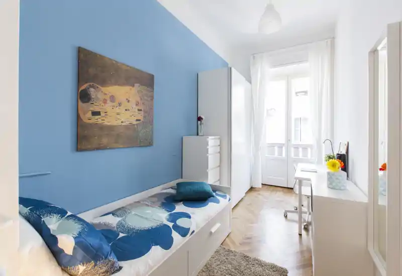 Bright single bedroom with a modern white bed, blue accent wall and natural light from a tall window — styled with cushions, a rug and simple storage for a cozy, fresh look.