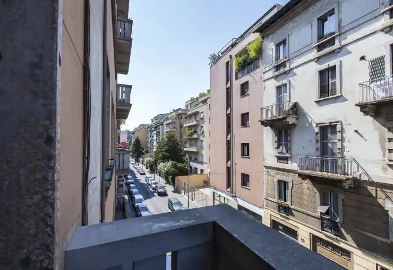 Vue depuis un balcon sur une rue résidentielle avec immeubles moyens et voitures garées; lumière du jour claire et végétation urbaine.