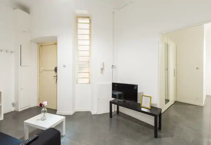 Salon lumineux et minimaliste avec murs blancs, une télévision sur un meuble noir, une table basse et un canapé compact. L'espace est propre, bien éclairé et meublé sobrement — idéal pour une location courte durée moderne.