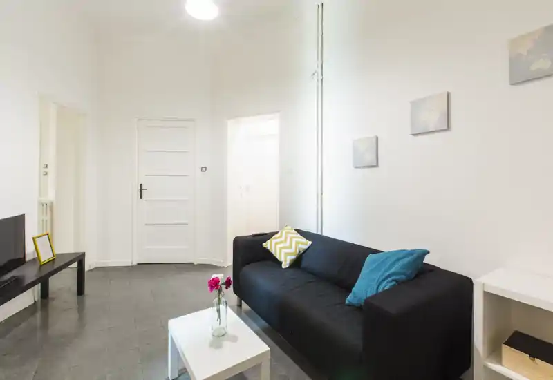 Petit salon moderne avec canapé noir, coussins décoratifs et une petite table basse blanche. Murs blancs lumineux et décoration minimaliste pour une atmosphère propre et accueillante.