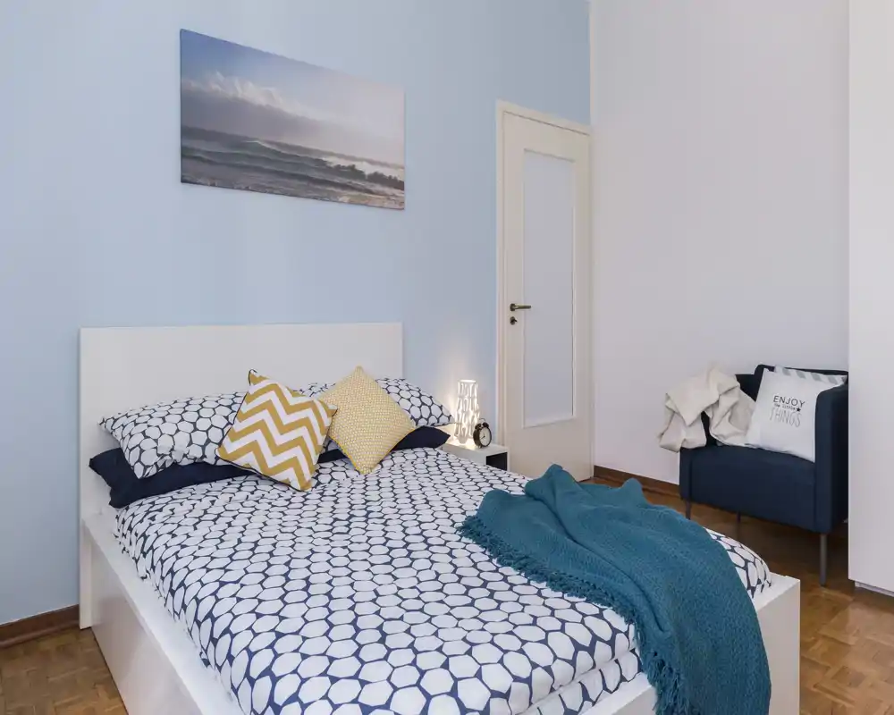 Dormitorio luminoso y bien presentado con cama doble estampada, manta acogedora y cojines decorativos, además de un pequeño sillón — perfecto para anuncios de alquiler.