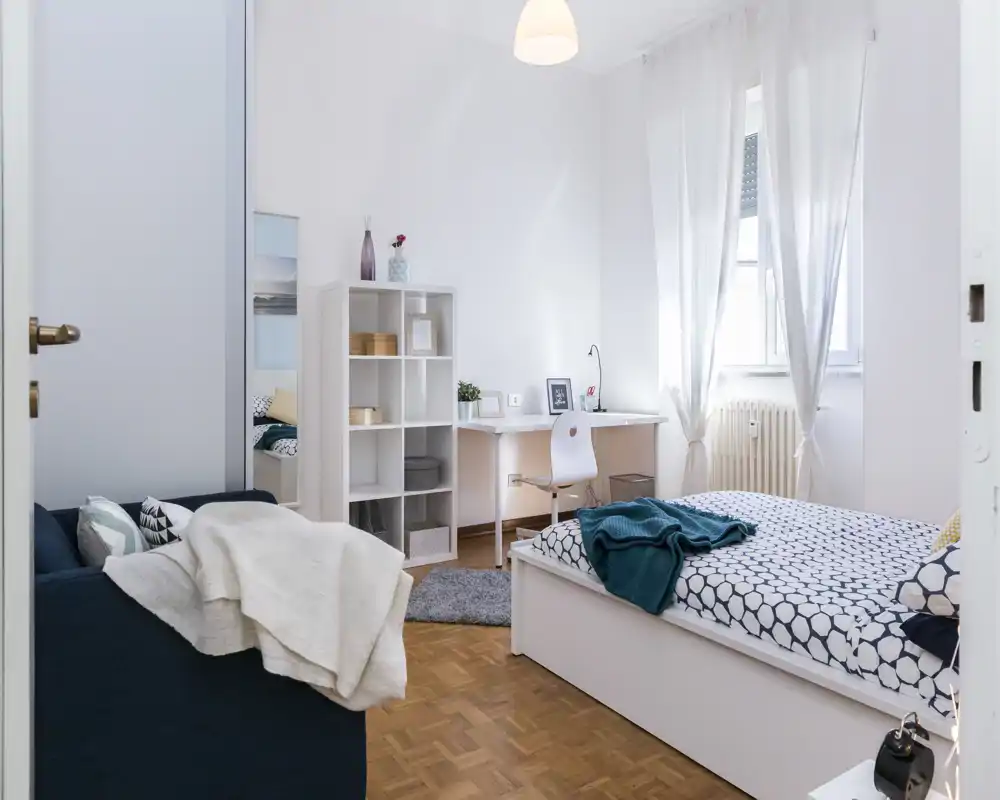 Dormitorio moderno y luminoso con cama doble cómoda, textiles acogedores y una pequeña zona de trabajo junto a la ventana. Paredes blancas y luz natural que crean una atmósfera amplia y atractiva para anuncios.
