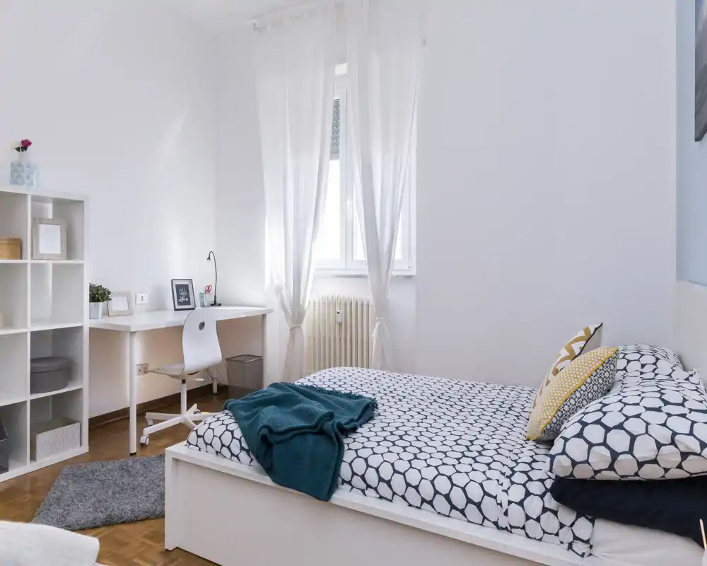 Dormitorio luminoso y moderno con cama doble con ropa de cama estampada, cojines decorativos y una manta turquesa. La luz natural y un escritorio sencillo crean un espacio cómodo y funcional.