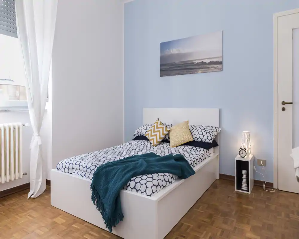Dormitorio luminoso y bien presentado con cama blanca de plataforma, ropa de cama estampada y manta acogedora; luz natural desde la ventana y lámpara de noche crean una atmósfera agradable.