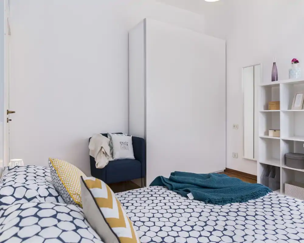 Dormitorio moderno y luminoso con ropa de cama estampada, cojines decorativos y una manta acogedora. Un gran armario y estanterías ofrecen almacenamiento; el espacio es aireado y bien decorado.
