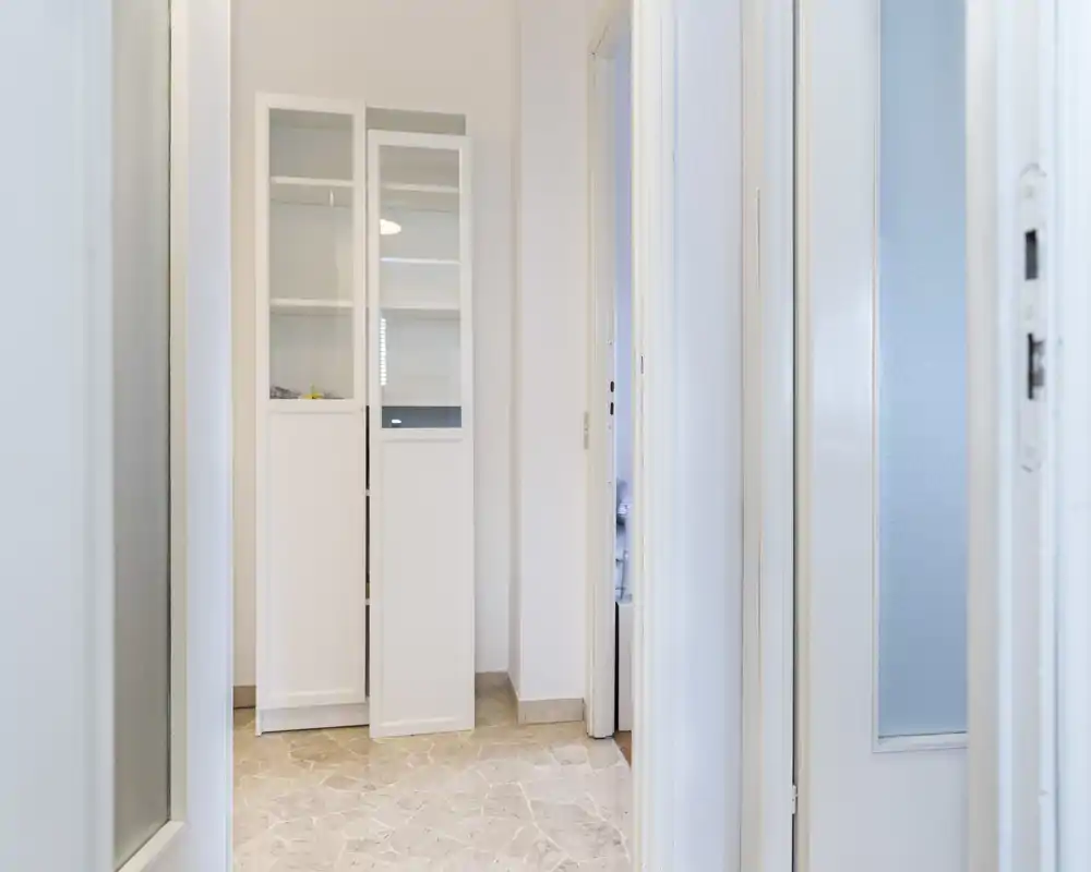 Entrada luminosa / pequeño pasillo con suelo de piedra clara y un armario alto blanco; decoración neutra y buena luz natural.