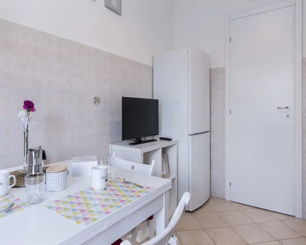 Esquina de cocina/comedor compacta con una mesa blanca pequeña preparada para café, un frigorífico y una TV sobre un mueble bajo; luminosa, ordenada y acogedora.