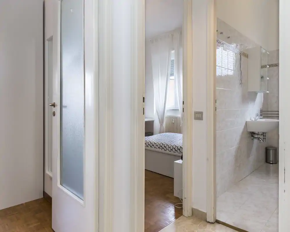 Toma luminosa desde el pasillo que muestra la entrada y la habitación con cama doble, ropa de cama estampada y luz natural de la ventana.