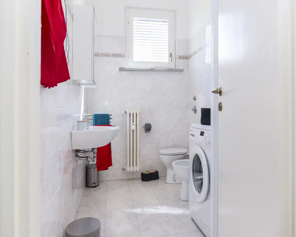 Baño luminoso con lavabo, inodoro, radiador y lavadora de carga frontal; azulejos blancos y una pequeña ventana que aporta luz natural.
