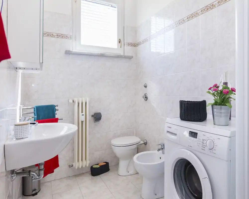 Baño limpio y luminoso con azulejos, lavabo, inodoro, bidé y lavadora, bien iluminado con luz natural.