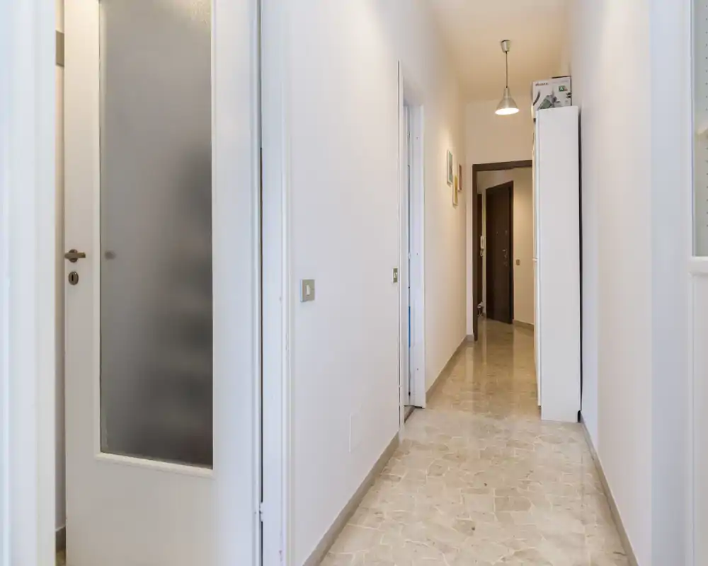 Pasillo de entrada luminoso con suelo de baldosas, paredes blancas limpias y un armario al fondo. La imagen destaca la longitud del pasillo y los accesos a las estancias.