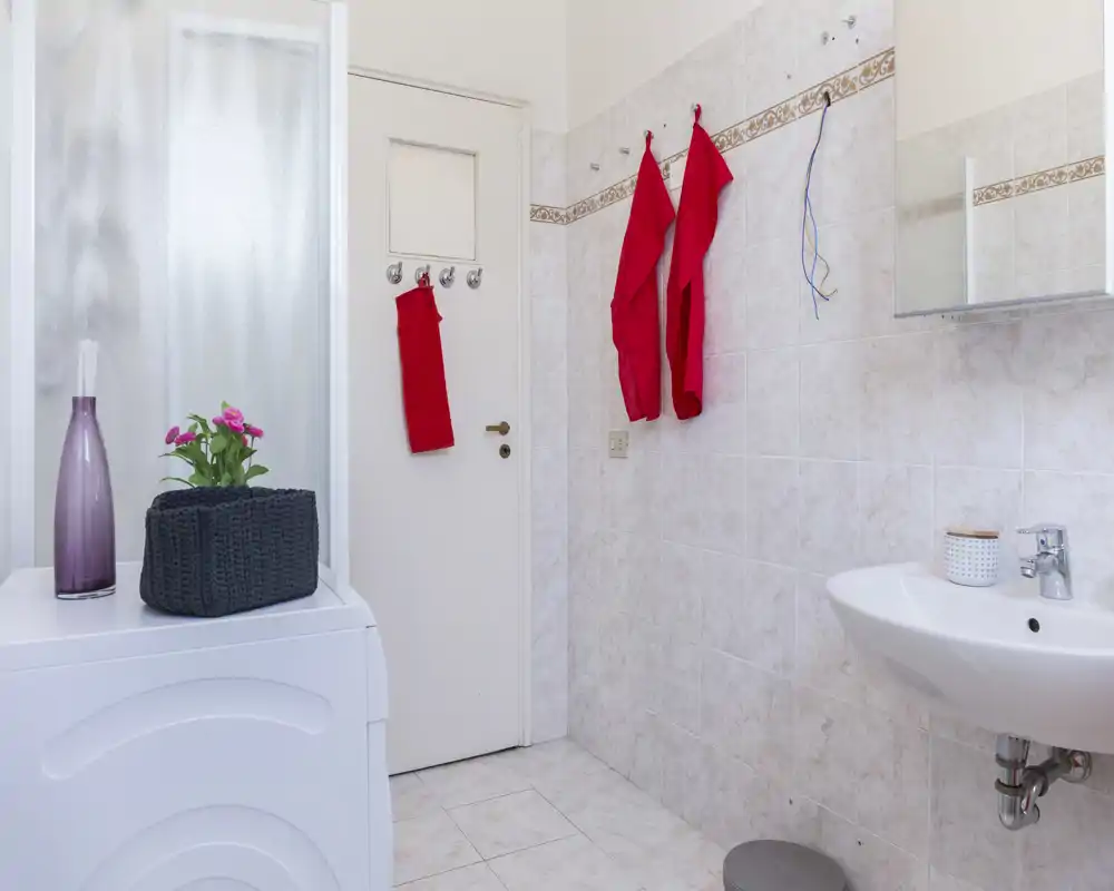 Baño alicatado y limpio con lavabo suspendido, armario con espejo y ganchos para toallas; luz brillante y decoración sencilla.