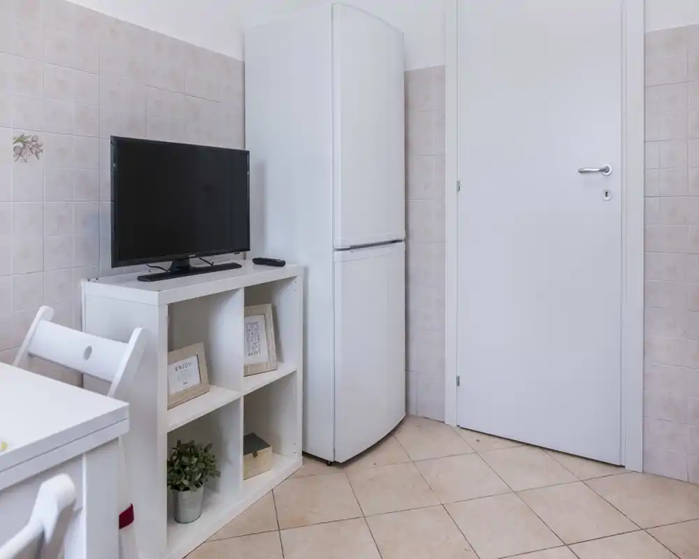 Rincón de cocina compacto con nevera, televisor sobre una estantería pequeña y una silla de comedor visible; azulejos y suelo claros mantienen el espacio luminoso y sencillo.