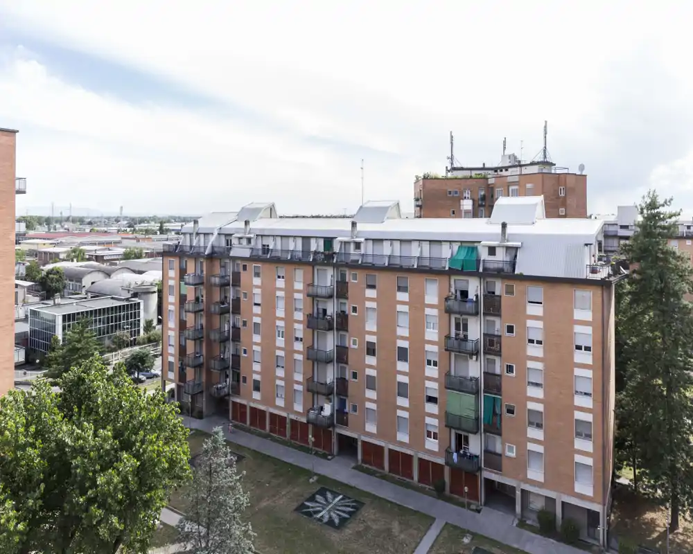 Vista exterior de un edificio residencial de varias plantas con balcones, una pequeña zona verde y árboles alrededor, tomada a plena luz del día.