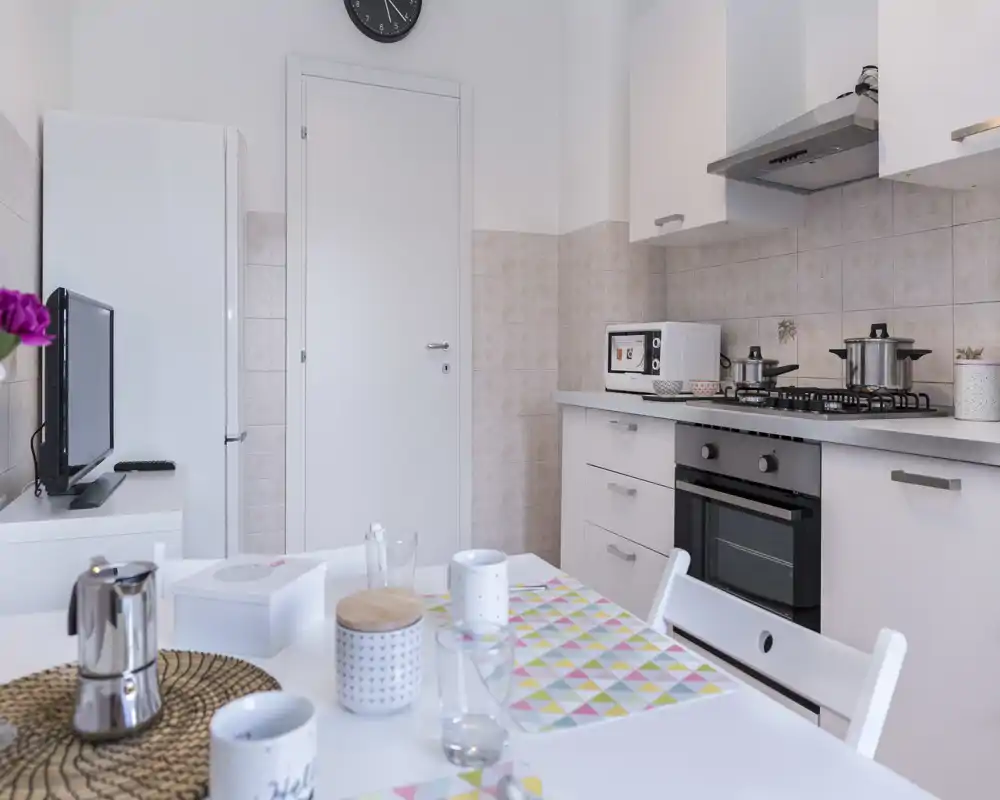 Cocina compacta y luminosa con cocina de gas, horno empotrado y una pequeña zona de comedor con mantelitos y tazas — ordenada, moderna y acogedora para el uso diario.