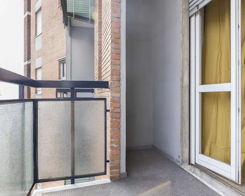 Pequeño balcón urbano con barandilla de vidrio esmerilado, pared de ladrillo y puerta corredera; apto para una silla o plantas.
