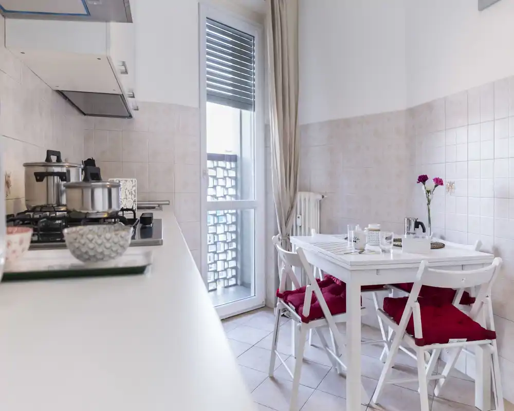 Cocina compacta y luminosa con mesa blanca y cojines rojos en las sillas; placa de gas con ollas y una puerta/ventana alta que aporta luz natural.