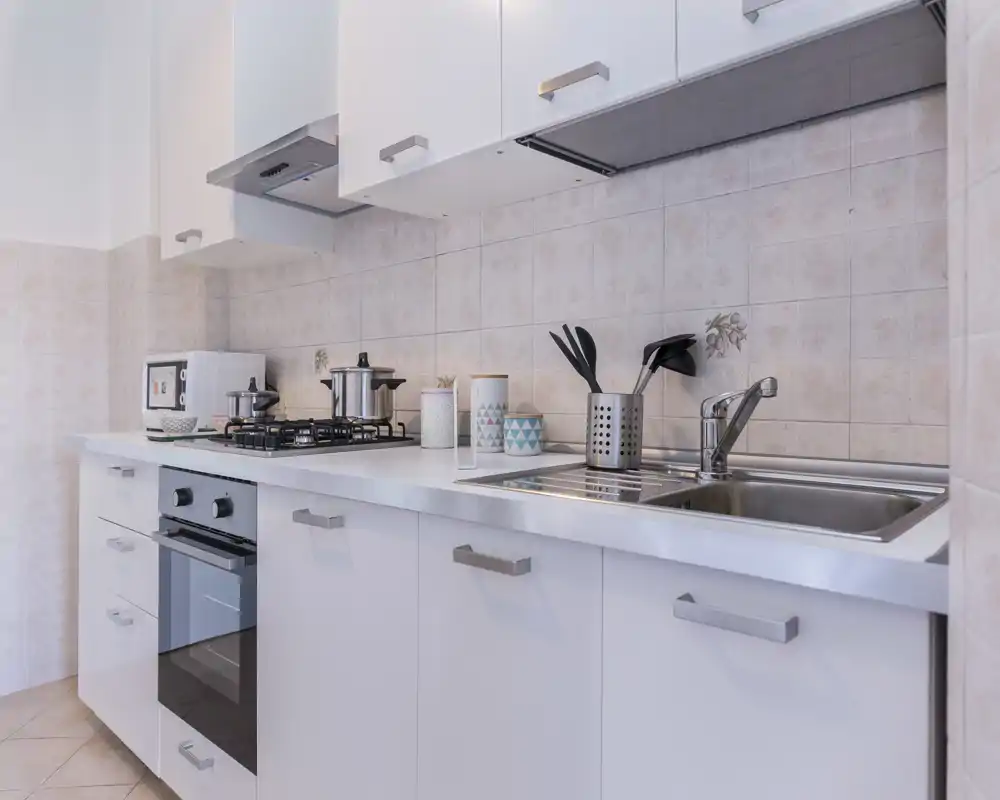 Cocina moderna y ordenada con armarios blancos, fregadero de acero, placa de gas y horno empotrado; botes y utensilios añaden un toque acogedor.