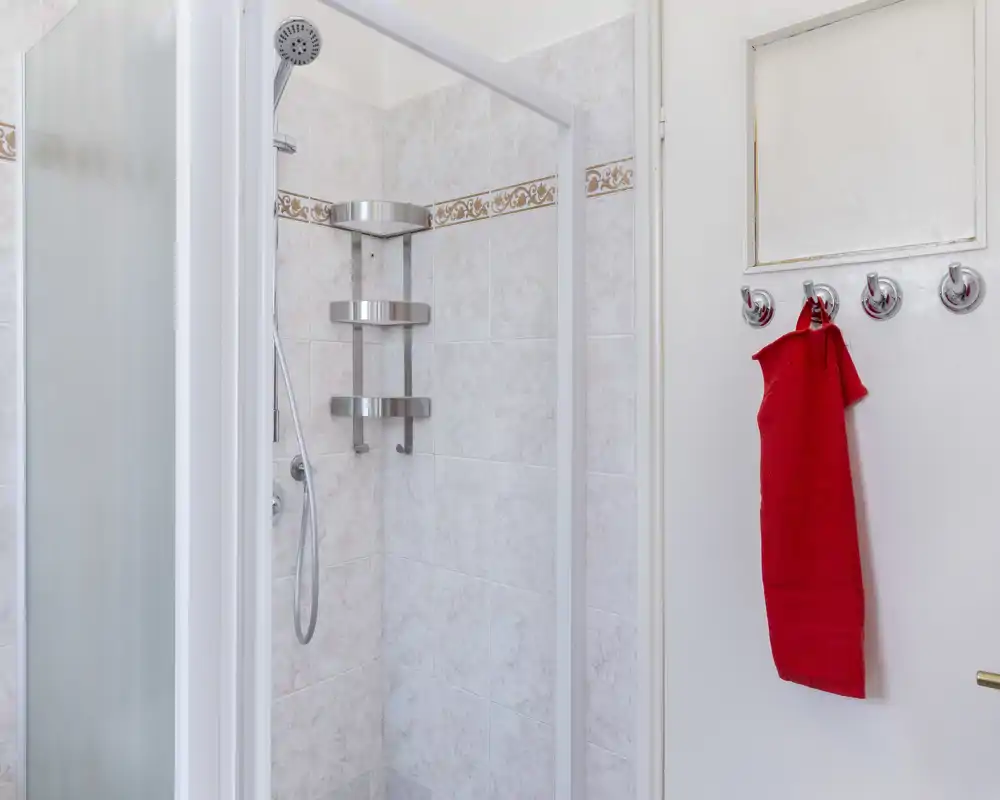 Baño empotrado con azulejos y mampara de ducha en la esquina, alcachofa de mano y estantería metálica; una toalla roja cuelga de los ganchos de la puerta.