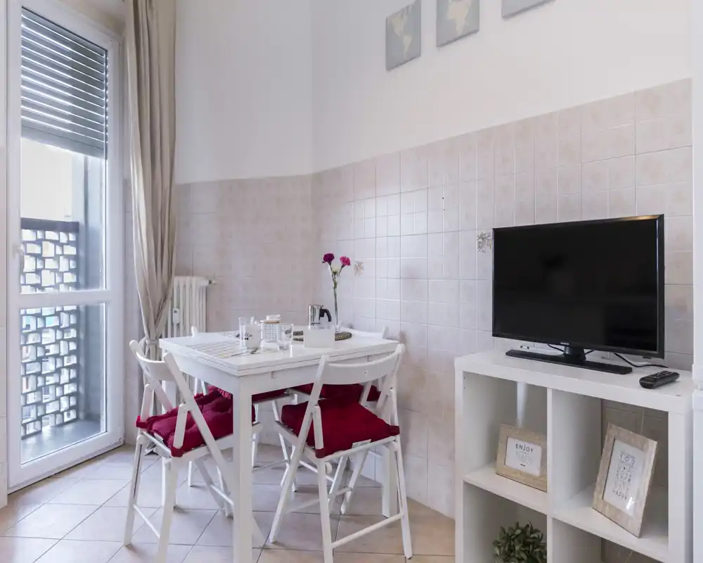 Acogedor rincón salón/comedor con mesa blanca pequeña, cuatro sillas con cojines rojos, TV en estantería baja y puerta de balcón que aporta luz natural.