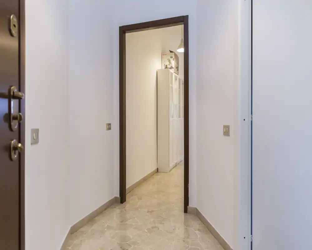 Entrada/pasillo limpio y estrecho con suelo de baldosas, paredes blancas y una puerta que conduce a otra habitación con muebles de almacenaje visibles.