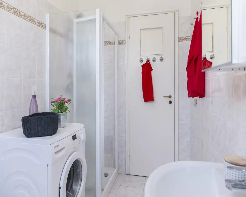Baño pequeño y bien iluminado con cabina de ducha, lavadora, lavabo y toallas rojas que aportan un toque de color.