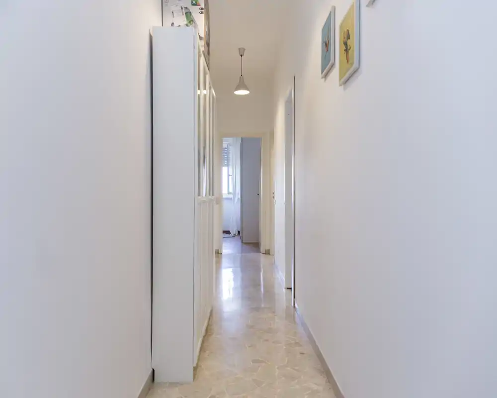 Pasillo de entrada luminoso y estrecho con paredes blancas, suelo de baldosas y lámpara colgante; decoración mínima y un mueble a la izquierda.