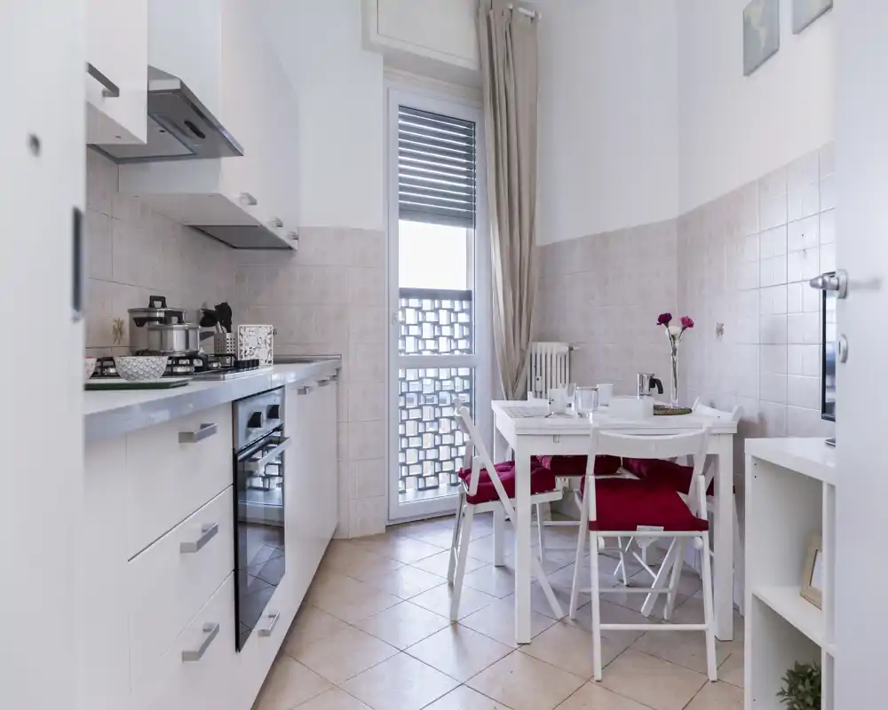 Cocina luminosa y compacta con horno integrado y una mesa pequeña junto a la puerta de cristal. Mobiliario blanco, suelo de baldosa y detalles decorativos que la hacen acogedora y funcional.