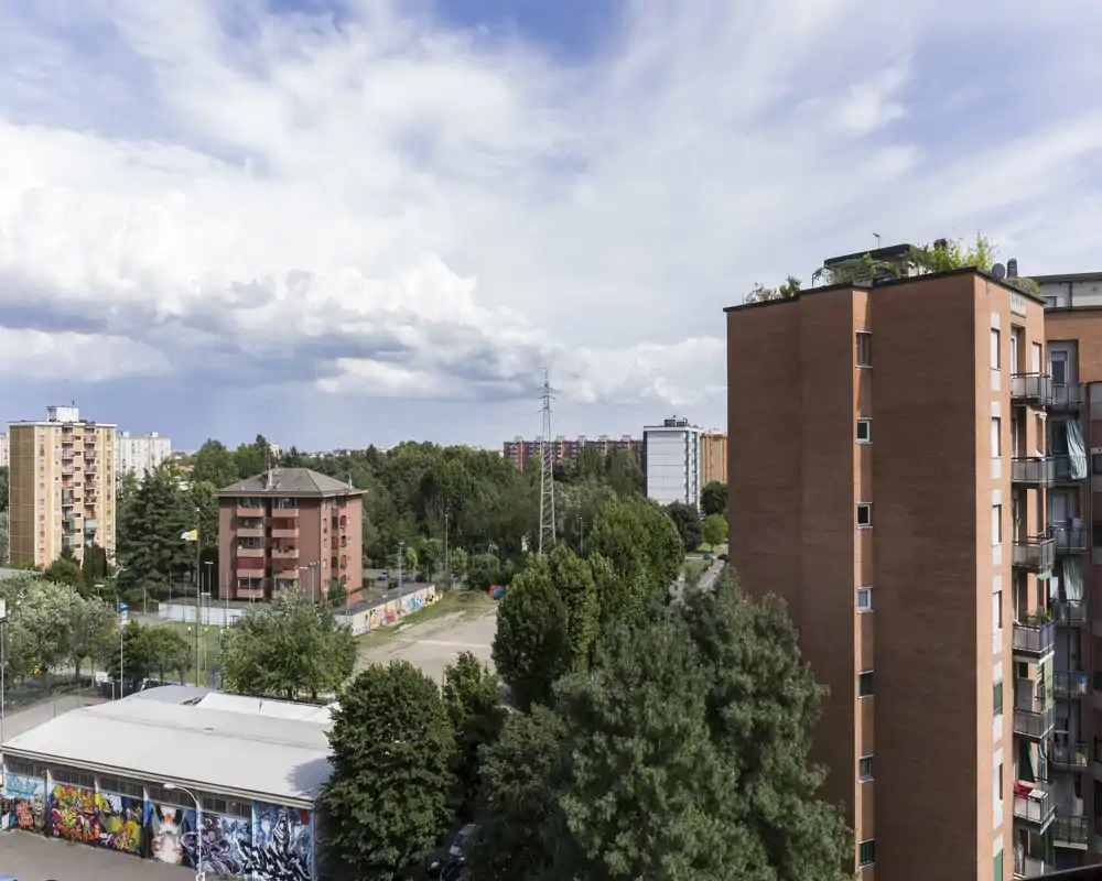 Vista urbana soleada desde un balcón/terraza con bloques de pisos, árboles y una pista deportiva—ideal para destacar el espacio exterior y las zonas verdes cercanas.