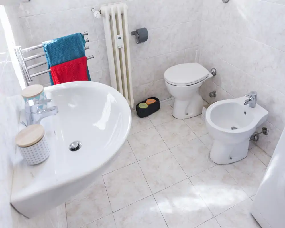 Baño limpio y luminoso con lavabo mural, inodoro y bidé sobre suelo de baldosas; toallas de colores en el radiador añaden un toque fresco.