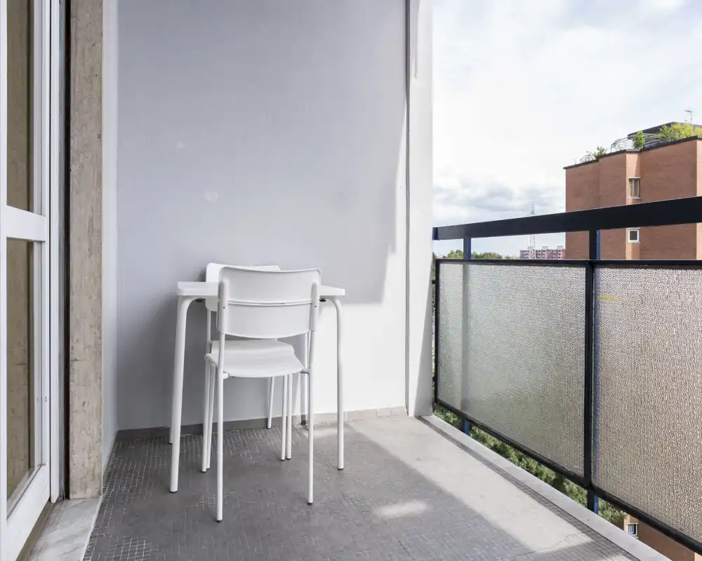 Balcón soleado con pequeña mesa blanca y sillas apiladas, barandilla de vidrio texturizado y agradable vista urbana — ideal para el café de la mañana.